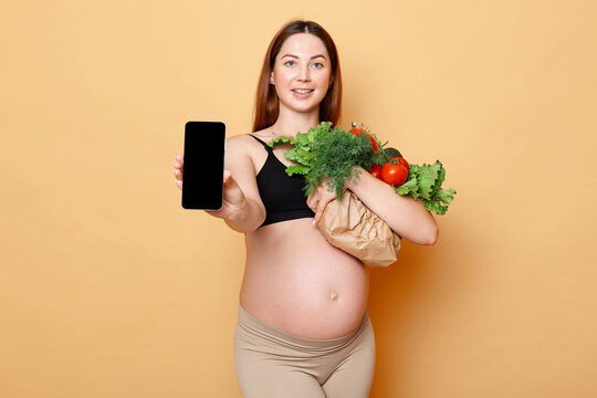 Winsome pregnant woman holding fresh vegetables posing isolated over beige background showing mobile phone with empty display empty space for advertisement.