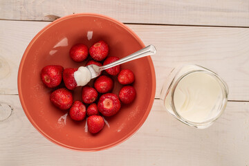 Ripe red strawberries with sugar and sour cream in a ceramic plate with a metal fork on a wooden table, macro, top view.