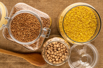 Dry cereals and seasonings in glass bowl with wooden spoons on jute cloth, macro, top view.