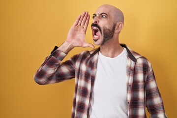 Hispanic man with beard standing over yellow background shouting and screaming loud to side with hand on mouth. communication concept.