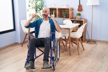Hispanic man with beard sitting on wheelchair holding new house keys smiling with an idea or question pointing finger with happy face, number one