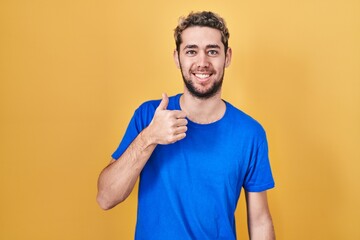 Hispanic man with beard standing over yellow background doing happy thumbs up gesture with hand. approving expression looking at the camera showing success.