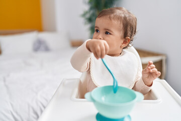 Fototapeta premium Adorable hispanic baby holding spoon sitting on highchair at bedroom