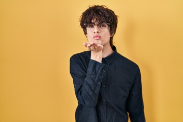 Young man wearing glasses over yellow background looking at the camera blowing a kiss with hand on...