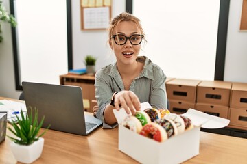 Young caucasian woman business worker using laptop holding doughnut at office
