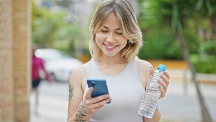 Young blonde woman using smartphone holding bottle of water at street