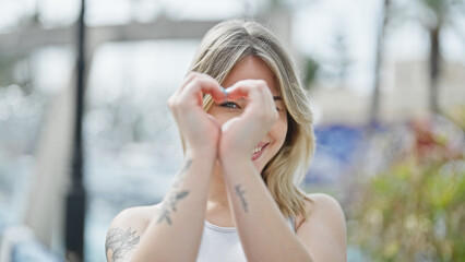 Young blonde woman smiling confident doing heart gesture with hands at park
