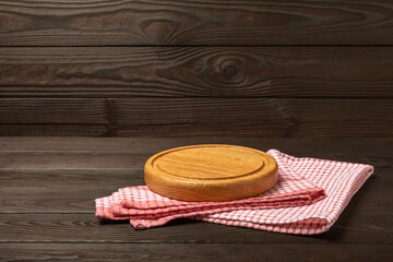 Empty wooden round pizza board on a kitchen brown wooden table. Cutting board with red checkered kitchen towel. Wooden pizza plate, copy space. Kitchenware. Recipe. Place for text. Flat lay.
