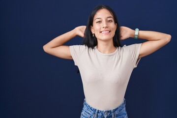 Young hispanic woman standing over blue background relaxing and stretching, arms and hands behind head and neck smiling happy