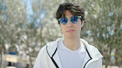 Young hispanic man standing with serious expression wearing sunglasses at park