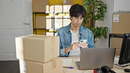 Young hispanic man ecommerce business worker using smartphone holding package at office