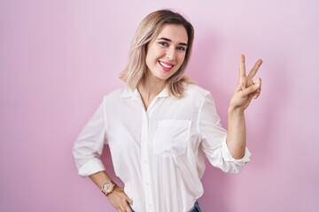 Young beautiful woman standing over pink background smiling looking to the camera showing fingers doing victory sign. number two.