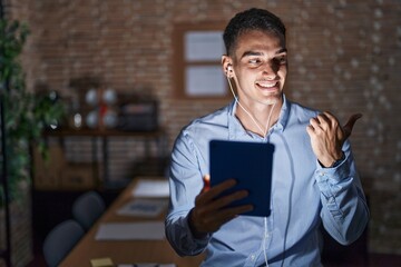 Handsome hispanic man working at the office at night smiling with happy face looking and pointing to the side with thumb up.