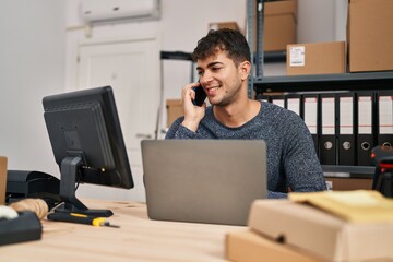 Young hispanic man ecommerce business worker using laptop talking on smartphone at office