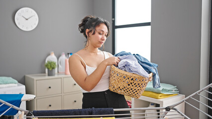 Young beautiful hispanic woman holding basket with clothes looking upset at laundry room