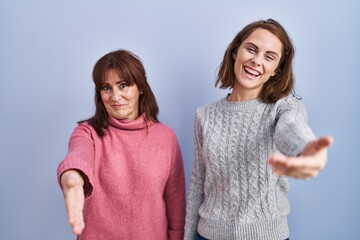 Mother and daughter standing over blue background smiling friendly offering handshake as greeting and welcoming. successful business.