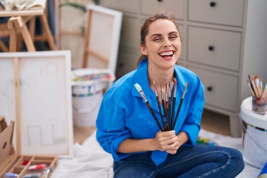 Young woman artist holding paintbrushes sitting on floor at art studio