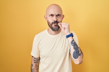 Hispanic man with tattoos standing over yellow background mouth and lips shut as zip with fingers....