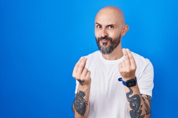 Hispanic man with tattoos standing over blue background doing money gesture with hands, asking for salary payment, millionaire business