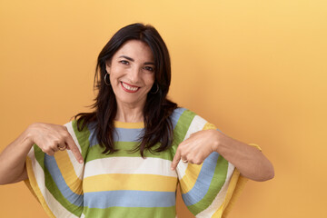 Middle age hispanic woman standing over yellow background looking confident with smile on face,...