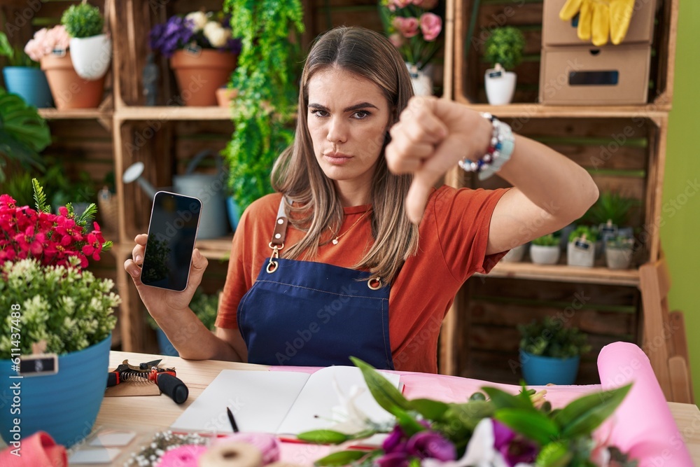 Sticker Hispanic young woman working at florist shop showing smartphone screen with angry face, negative sign showing dislike with thumbs down, rejection concept