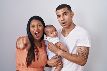 Young hispanic couple with baby standing together over isolated background pointing with finger surprised ahead, open mouth amazed expression, something on the front