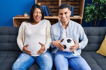 Young hispanic couple expecting a baby sitting on the sofa holding ball and tummy winking looking...