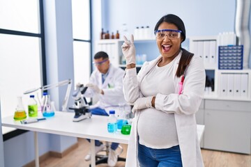 Young hispanic woman expecting a baby working at scientist laboratory smiling with happy face...