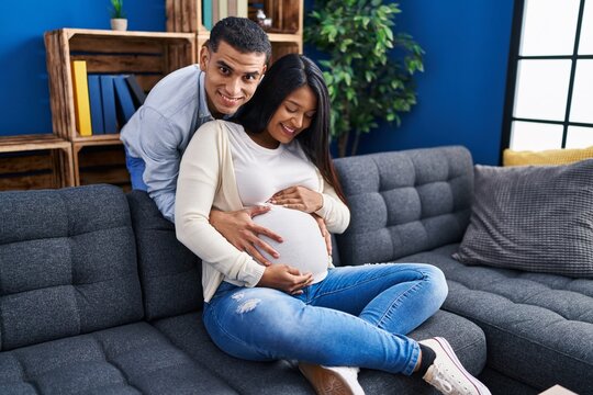 Young latin couple expecting baby hugging each other sitting on sofa at home