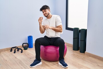 Hispanic man with beard sitting on pilate balls at yoga room disgusted expression, displeased and...