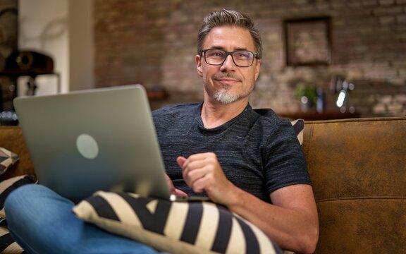 Older man working online with laptop computer at home sitting on couch in living room. Home office, browsing internet. Portrait of happy, mature age, middle age, mid adult man in 50s, smiling.