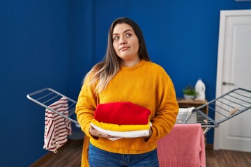 Young hispanic woman holding clean and folded laundry smiling looking to the side and staring away thinking.