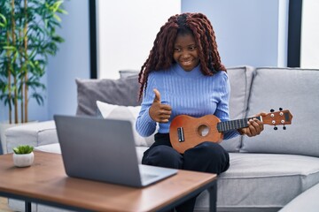 African woman playing ukulele at home doing video call smiling happy and positive, thumb up doing excellent and approval sign