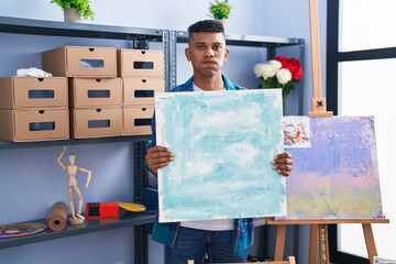 Young hispanic man holding canvas at artist studio puffing cheeks with funny face. mouth inflated...