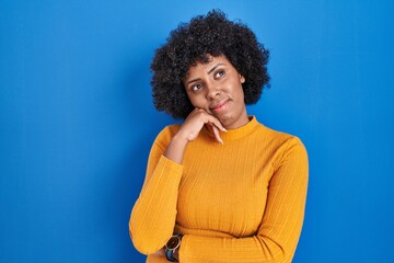 Fototapeta na wymiar Black woman with curly hair standing over blue background with hand on chin thinking about question, pensive expression. smiling with thoughtful face. doubt concept.