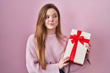 Young caucasian woman holding gift smiling looking to the side and staring away thinking.