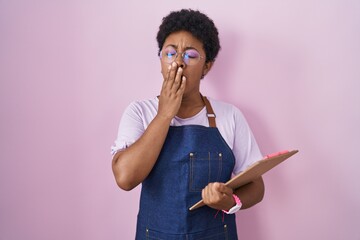 Young african american woman wearing professional waitress apron holding clipboard bored yawning...