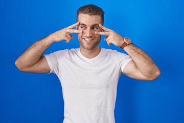Naklejka premium Young caucasian man standing over blue background doing peace symbol with fingers over face, smiling cheerful showing victory