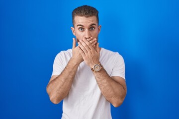 Young caucasian man standing over blue background shocked covering mouth with hands for mistake....