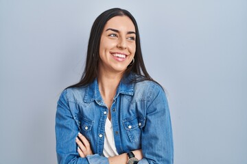 Hispanic woman standing over blue background looking away to side with smile on face, natural...