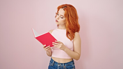 Young redhead woman reading book with serious expression over isolated pink background