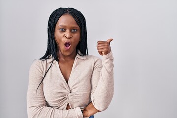 African woman with braids standing over white background surprised pointing with hand finger to the side, open mouth amazed expression.