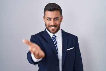 Handsome hispanic man wearing suit and tie smiling cheerful offering palm hand giving assistance and acceptance.