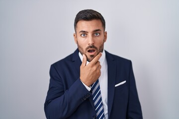 Handsome hispanic man wearing suit and tie looking fascinated with disbelief, surprise and amazed expression with hands on chin