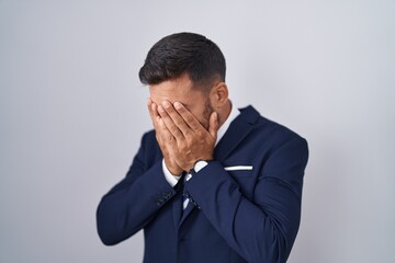 Handsome hispanic man wearing suit and tie with sad expression covering face with hands while...