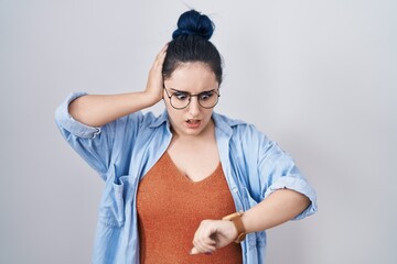 Young modern girl with blue hair standing over white background looking at the watch time worried,...