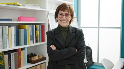 Mature hispanic woman teacher standing by white board with crossed arms at library university