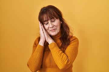 Middle age hispanic woman standing over yellow background sleeping tired dreaming and posing with hands together while smiling with closed eyes.