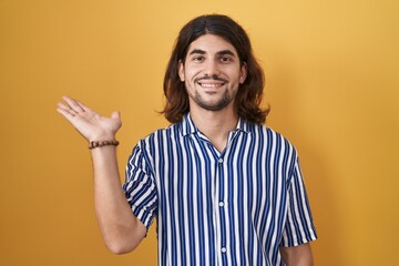 Hispanic man with long hair standing over yellow background smiling cheerful presenting and pointing with palm of hand looking at the camera.