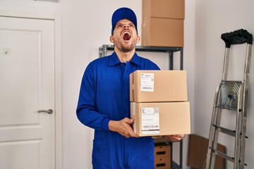 Young hispanic man working on moving service holding boxes angry and mad screaming frustrated and furious, shouting with anger looking up.